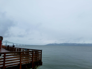 Wet pier to the left, with bridge in distance. Pier is overlooking large body of water with mountains in the background.